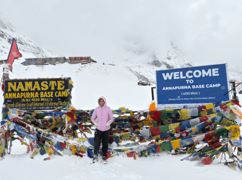 Angira Shah stands alone at Annapurna Base Camp in front of prayer flags and snow-covered peaks.