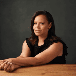 Portrait of CBS News journalist Anne-Marie Green seated at a wooden table, wearing a sleeveless black top, with a calm and confident expression against a dark background.