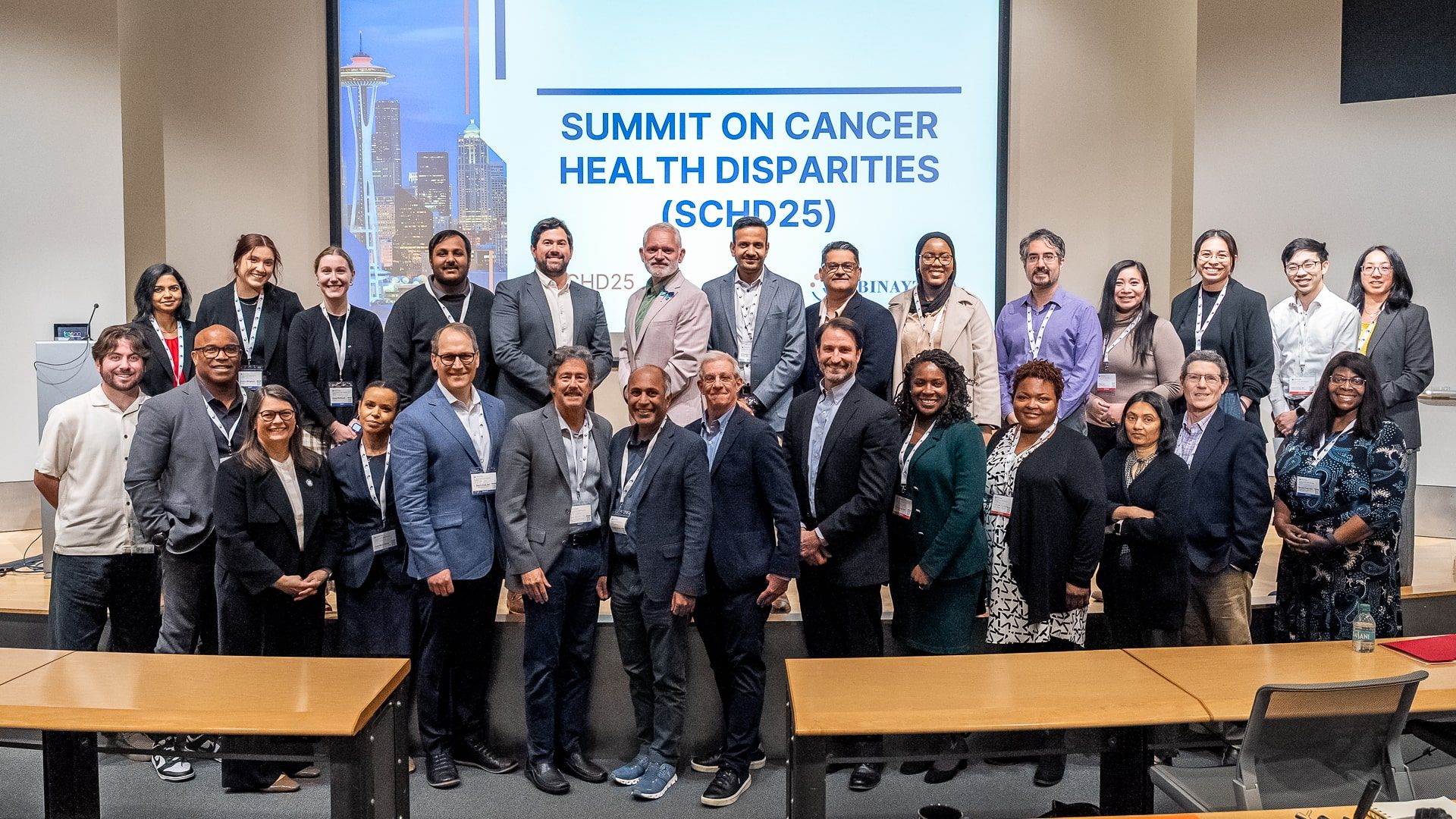 Group photo of SCHD25 faculty and participants at the Day 3 finale of the Summit on Cancer Health Disparities, with the Seattle skyline and SCHD25 signage in the background, highlighting global oncology leaders advancing cancer health equity.