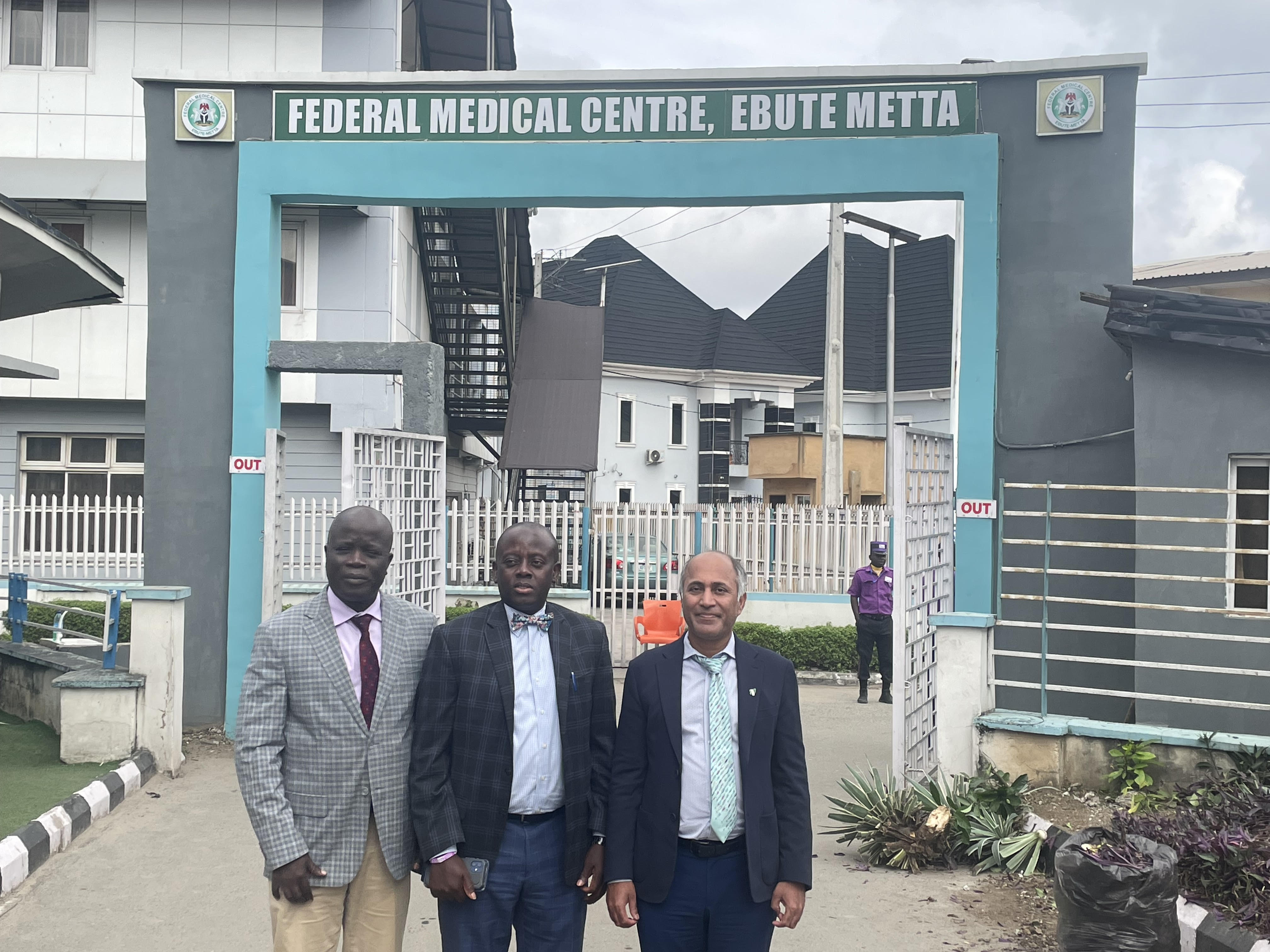 Dr. Rasaq Olaosebikan, Dr. Daniel Ezekwudo, and Dr. Binay Shah standing at the entrance of Federal Medical Center Ebute Metta in Lagos, Nigeria.