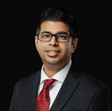 Dr. Kartik Anand in a black suit, white shirt, and red tie, smiling against a black background.