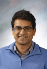 Dr. Vishal Vashistha, wearing a blue sweater and glasses, smiling in front of a soft gradient background.